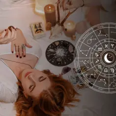 Kobieta z rudymi włosami leży na łóżku, trzymając w dłoni kamień. Obok niej talia kart, świece i koło zodiaku. 19 października znak zodiaku to Waga.