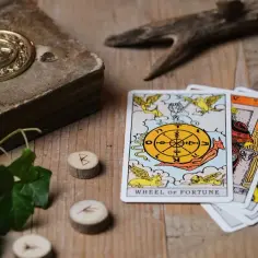 Tarotowe karty, w tym Koło Fortuny, leżą obok starej księgi i run. Czekasz na horoskop tygodniowy miłosny?